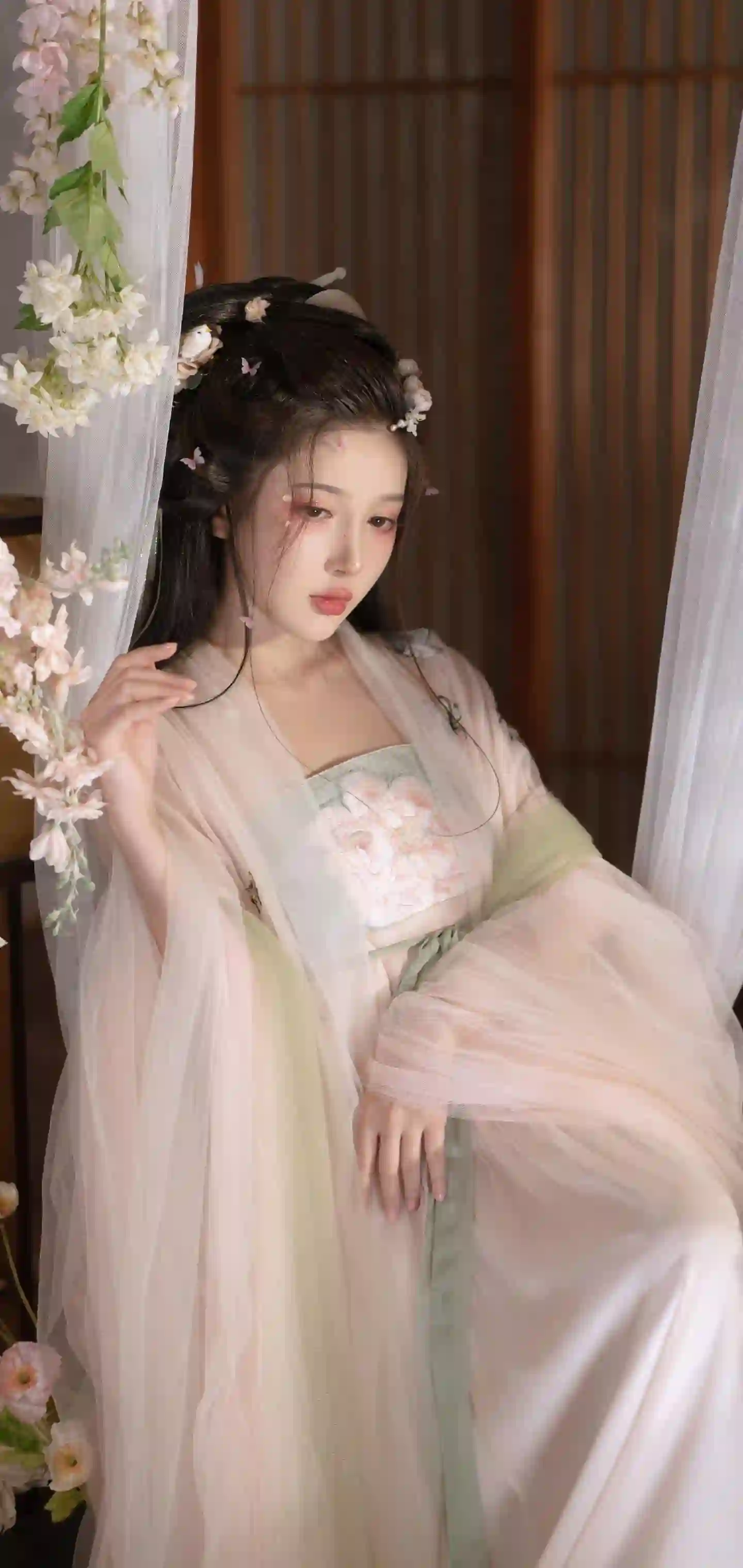 一位穿着浅色汉服的年轻女性 头戴花朵装饰 站在花枝和帘子旁 营造出古典优雅的氛围