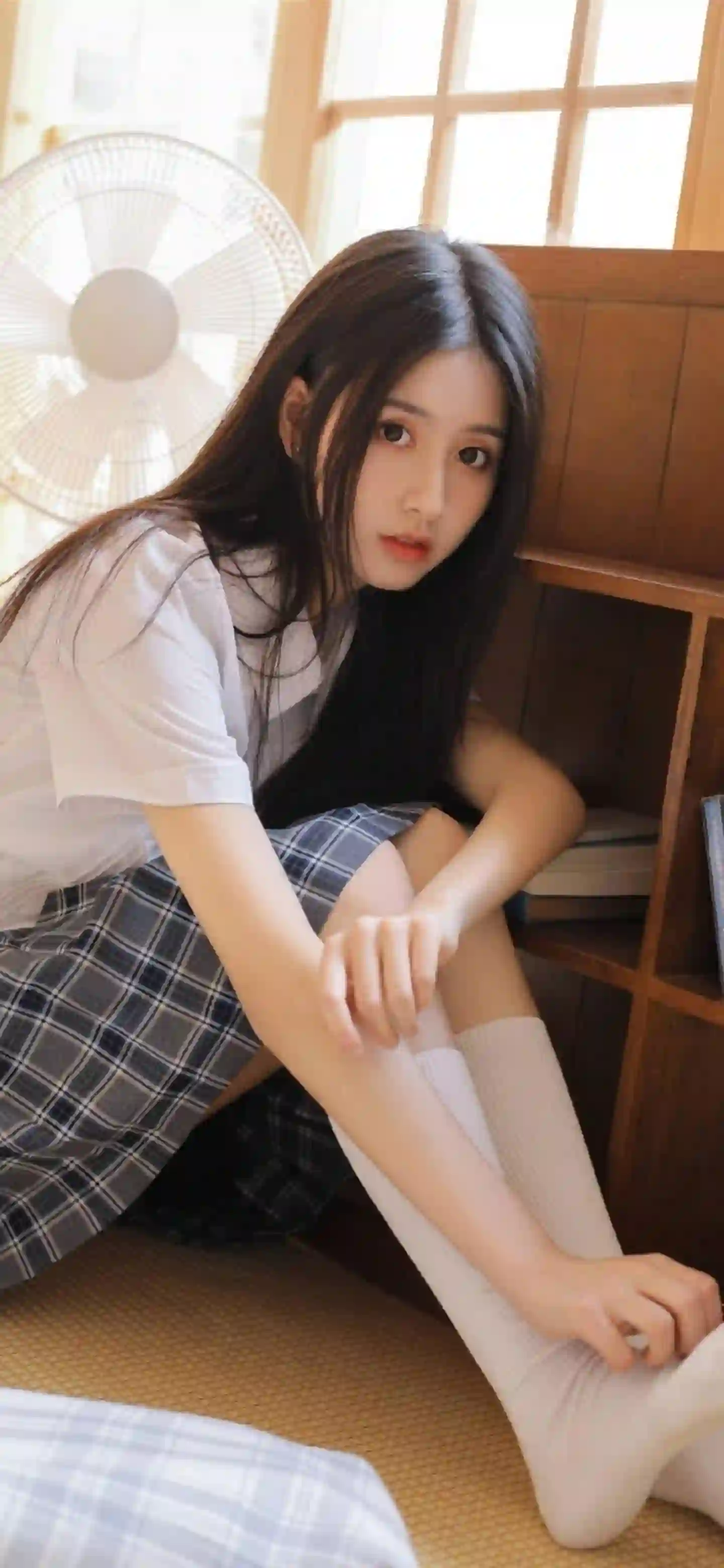 图片展示了一个长发女孩，穿着白色短袖T恤和蓝灰格子裙，搭配白色长袜，坐在木质地板上，背景有电风扇和书架，氛围清新自然。