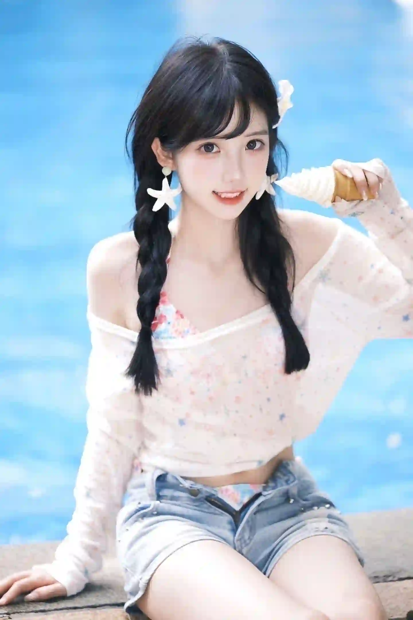 一位扎着双马尾的年轻女孩，身穿白色花纹露肩上衣和牛仔短裤，手持冰淇淋，坐在海边石台上，背景是蓝色的大海。