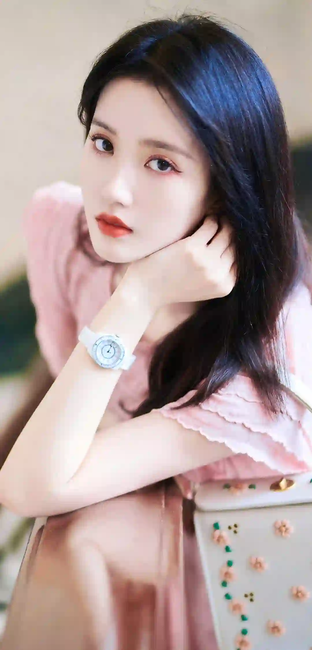 一位穿着粉色连衣裙的年轻女性 手托下巴 佩戴白色手表 黑色长发披肩 背景模糊 展现出温柔优雅的气质