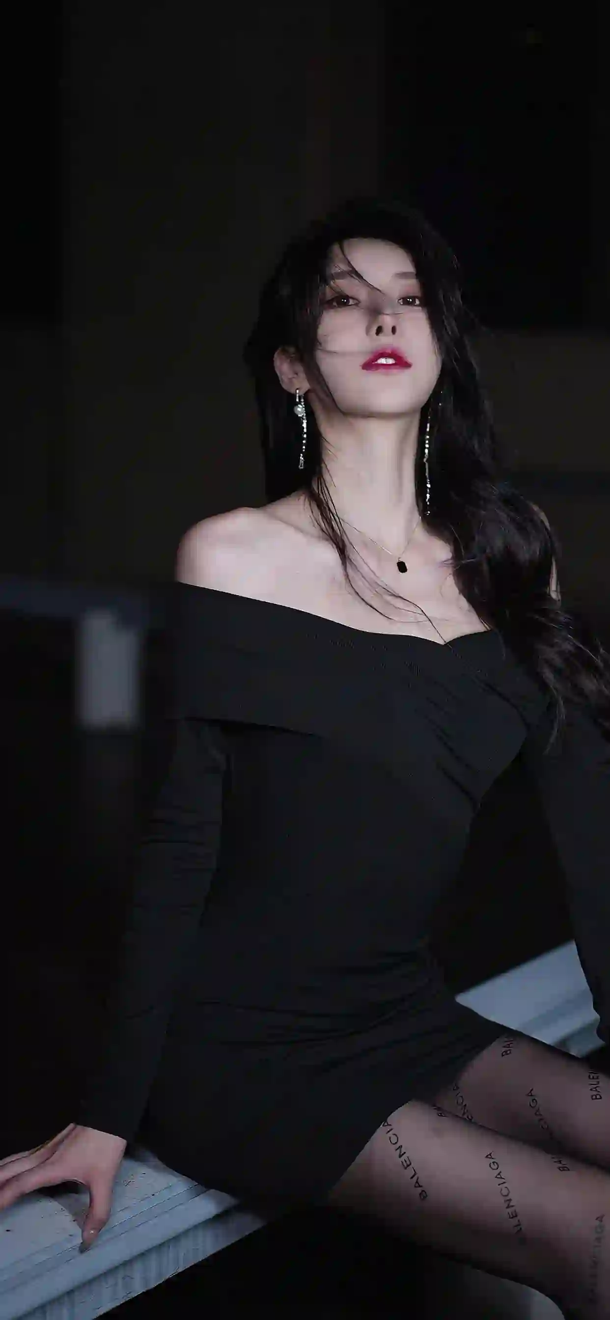 一位穿着黑色露肩连衣裙的女性 长发披肩 红唇艳丽 坐在白色台面上 背景深色 营造出时尚优雅的氛围