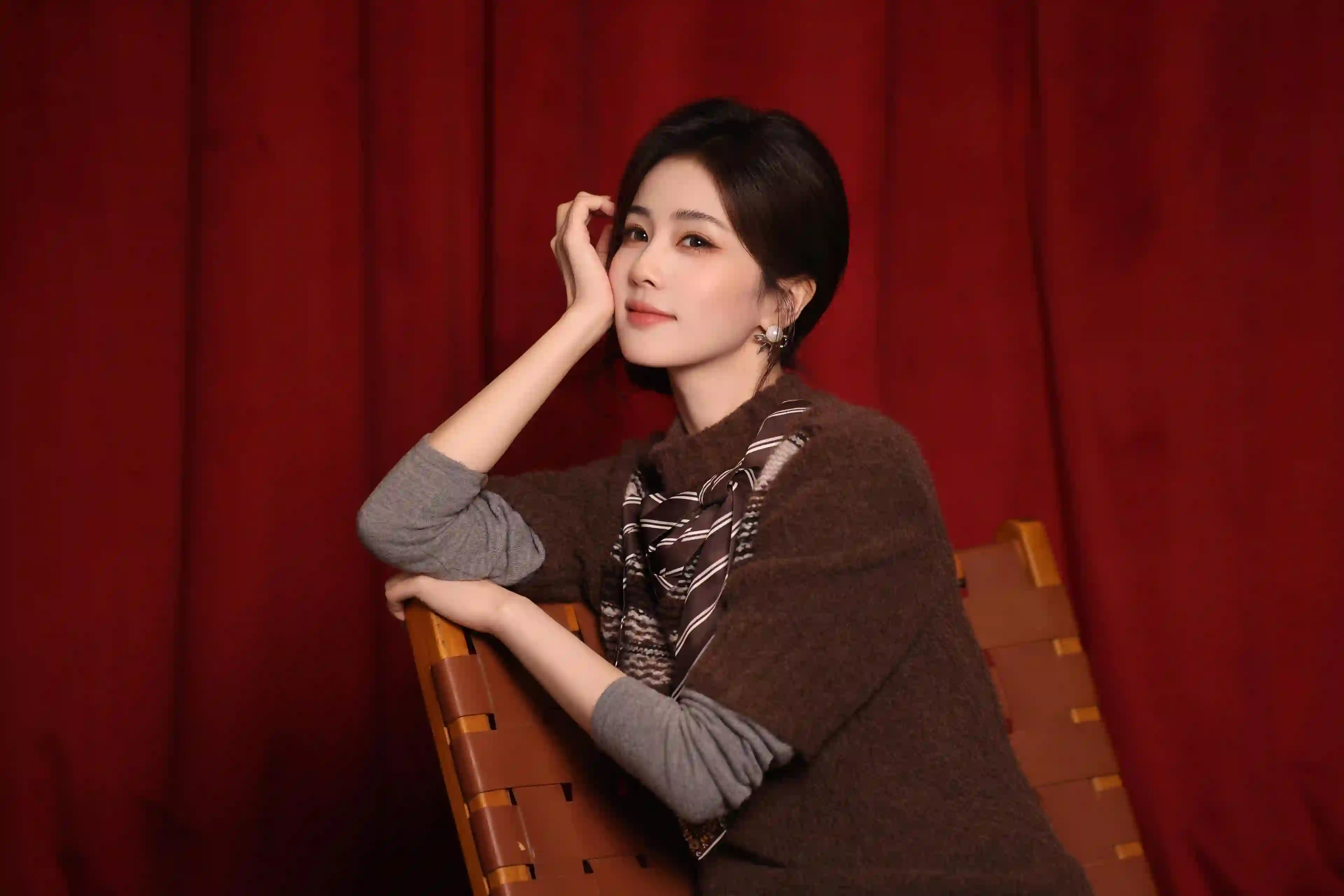 一位穿着棕色斗篷和条纹丝巾的女性 坐在椅子上手托脸颊 背景为红色幕布