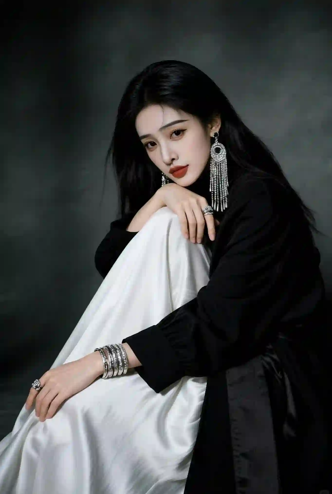 一位长发女性穿着黑色上衣和白色裙子，佩戴银色耳环、手镯和戒指，背景深色，姿态优雅时尚。