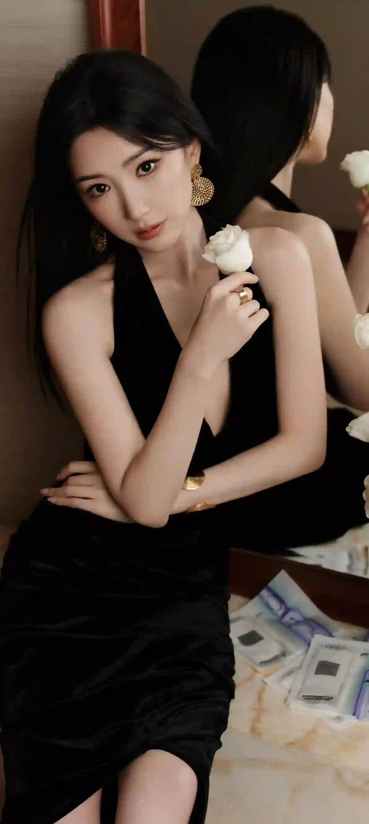 图片展示了一位穿着黑色吊带裙的优雅女性 她手持一朵白玫瑰 佩戴金色大耳环和手镯 背景是镜子反射出她的背影 整体氛围高级且充满艺术感