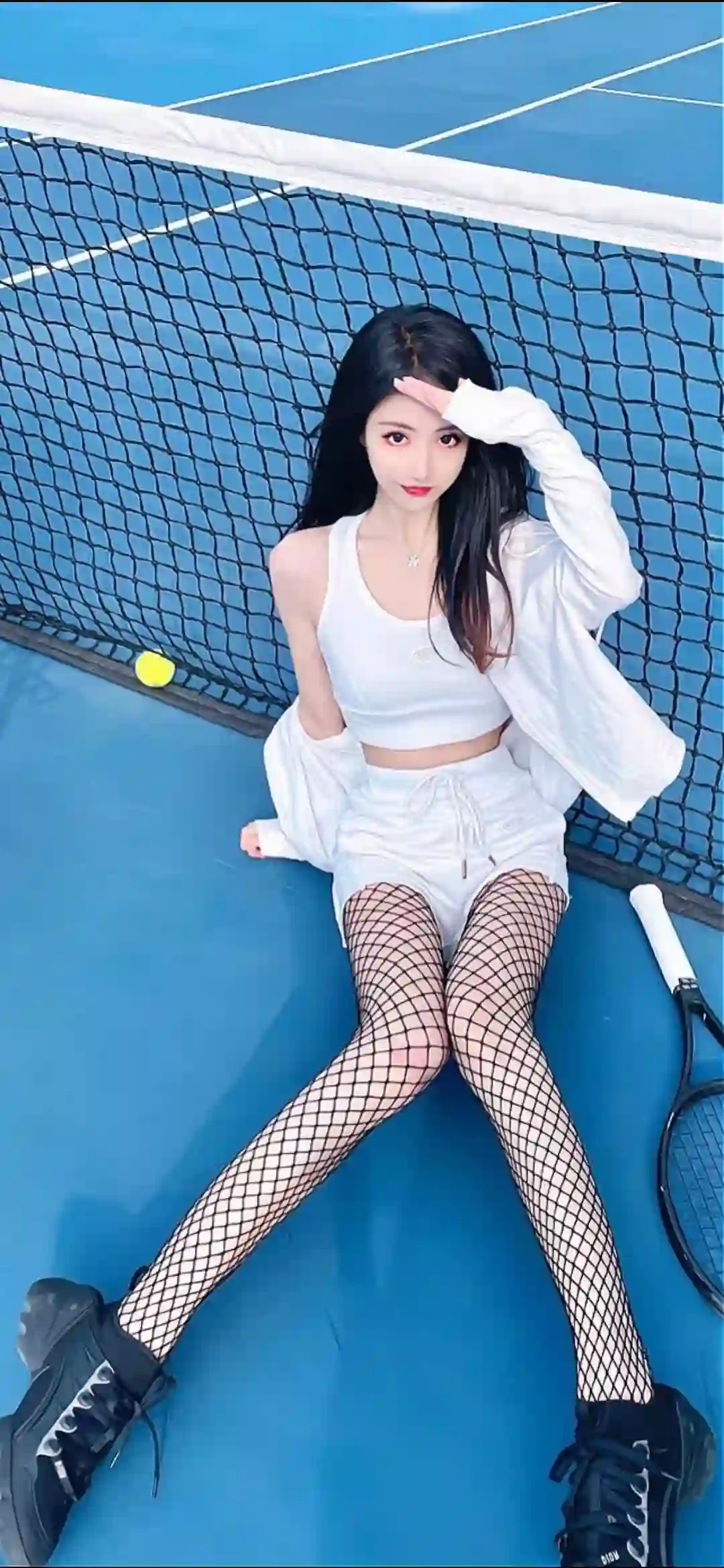 一位长发女性坐在蓝色网球场上 穿着白色背心 短裤和外套 搭配黑色网袜和运动鞋 手持网球拍 姿态优雅自信
