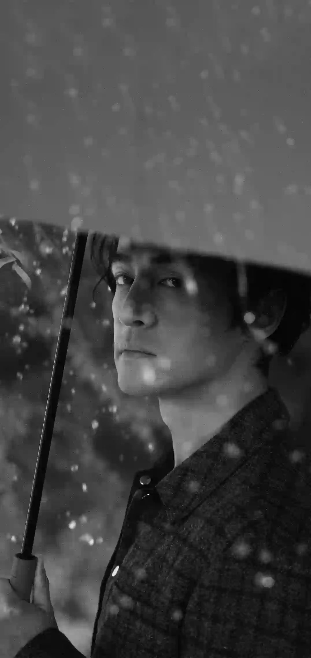 一张黑白摄影图片 展示一位男子在雨中撑伞 表情沉思 背景雨滴点点 营造忧郁艺术氛围