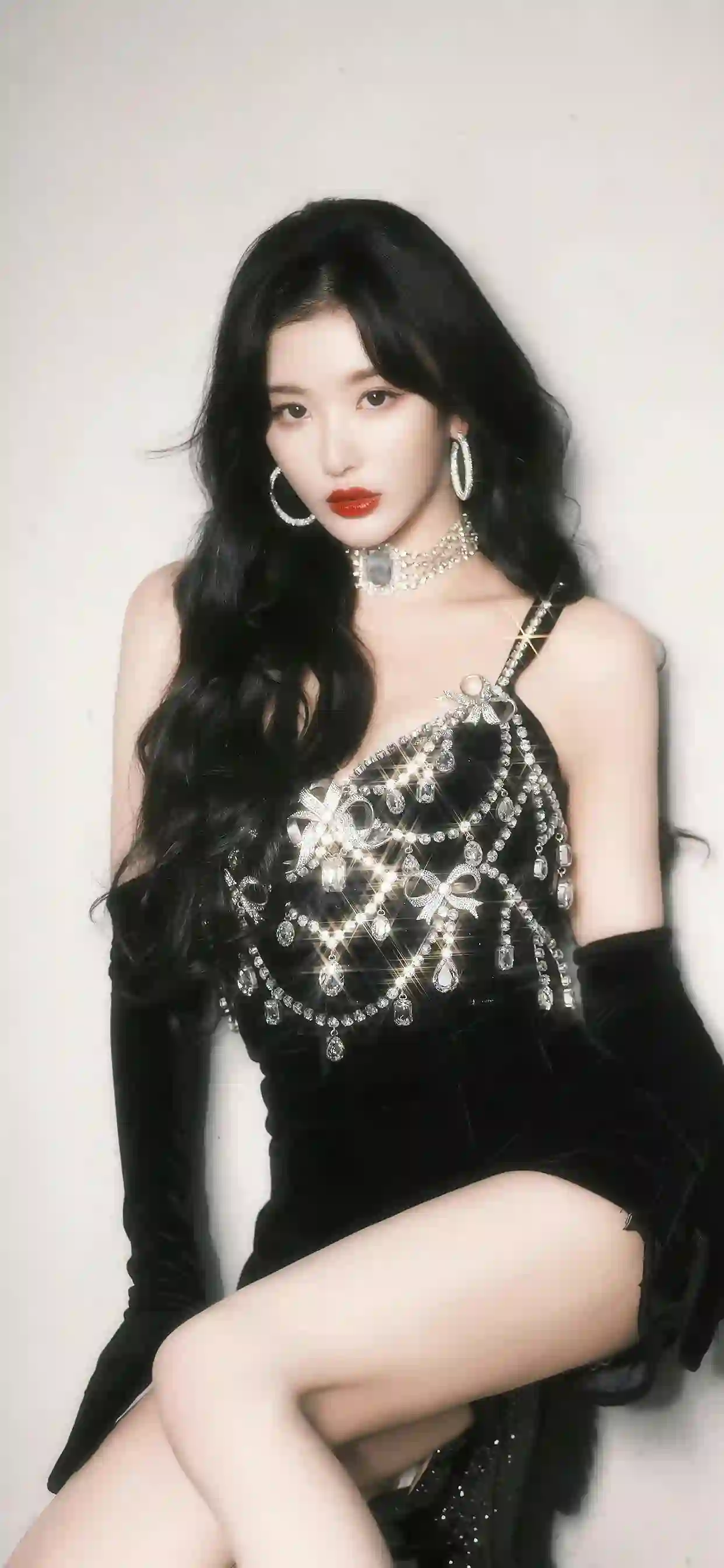 一位黑发长卷发女性穿着黑色亮片连衣裙 佩戴银色耳环和项链 戴长手套 坐姿优雅 红唇妆容 背景为浅色 突出性感时尚感