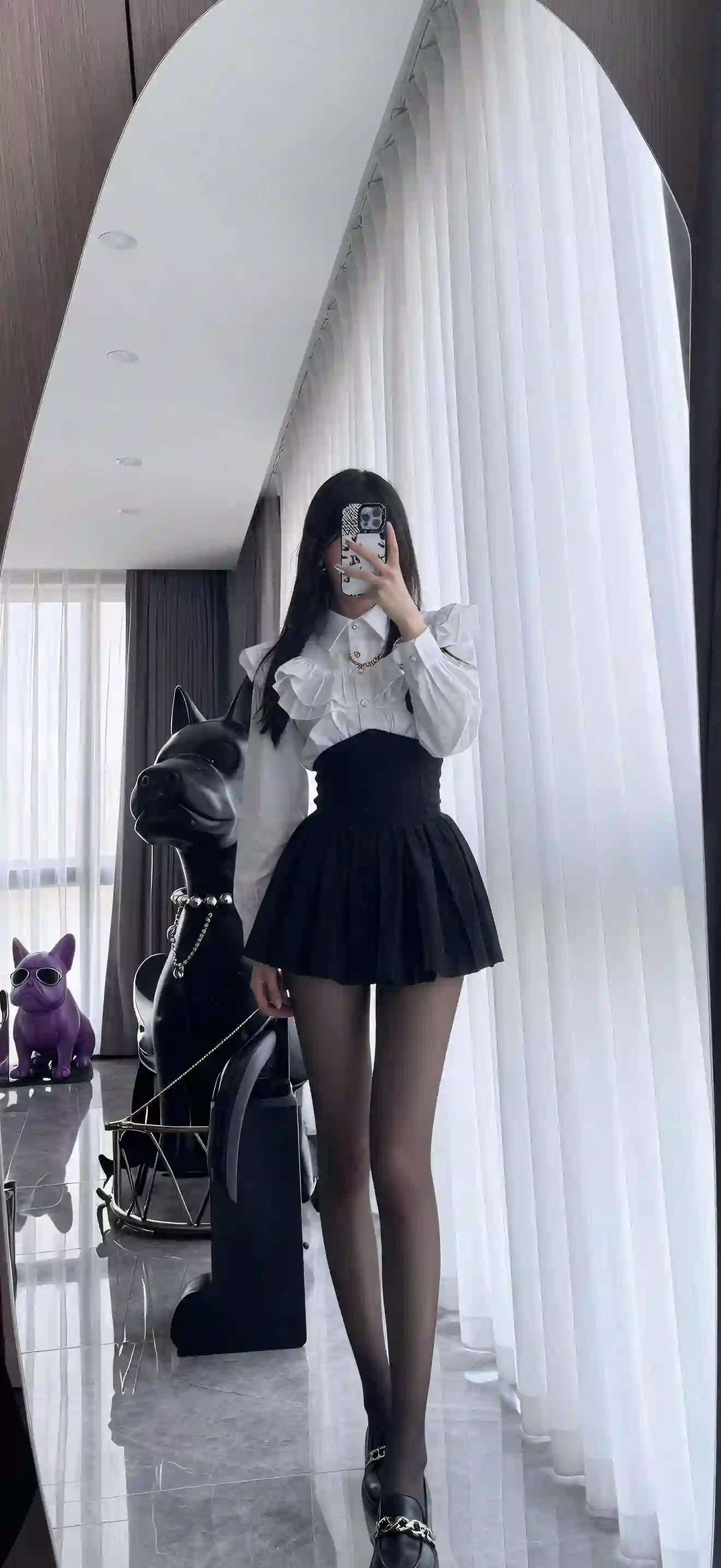 长发女孩在室内手持手机自拍，身穿白色荷叶边衬衫和黑色百褶短裙，搭配黑色丝袜和黑色鞋子，背景有白色窗帘和装饰性狗雕塑。