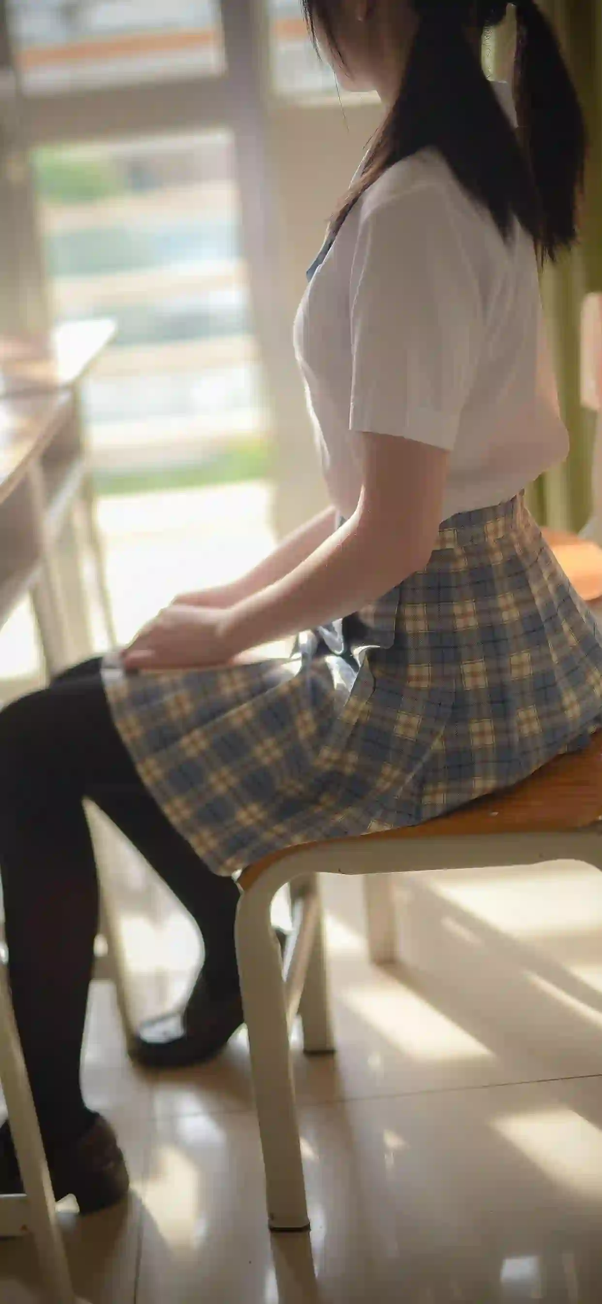 一个女孩坐在教室椅子上 穿着白色T恤和蓝黄格子裙 黑色丝袜 阳光从窗户照入 营造温暖逆光氛围 整体呈现校园青春感