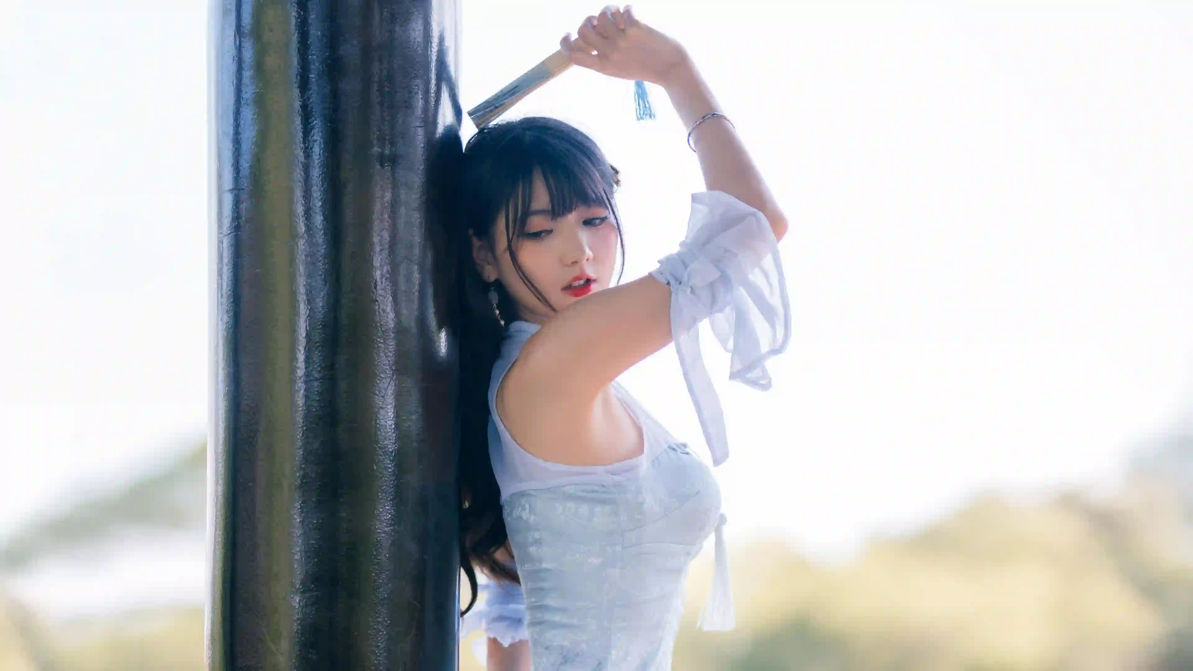 一位穿着淡蓝色汉服的年轻女性，手持折扇倚靠在黑色柱子上，背景是明亮的户外自然光，氛围梦幻唯美。