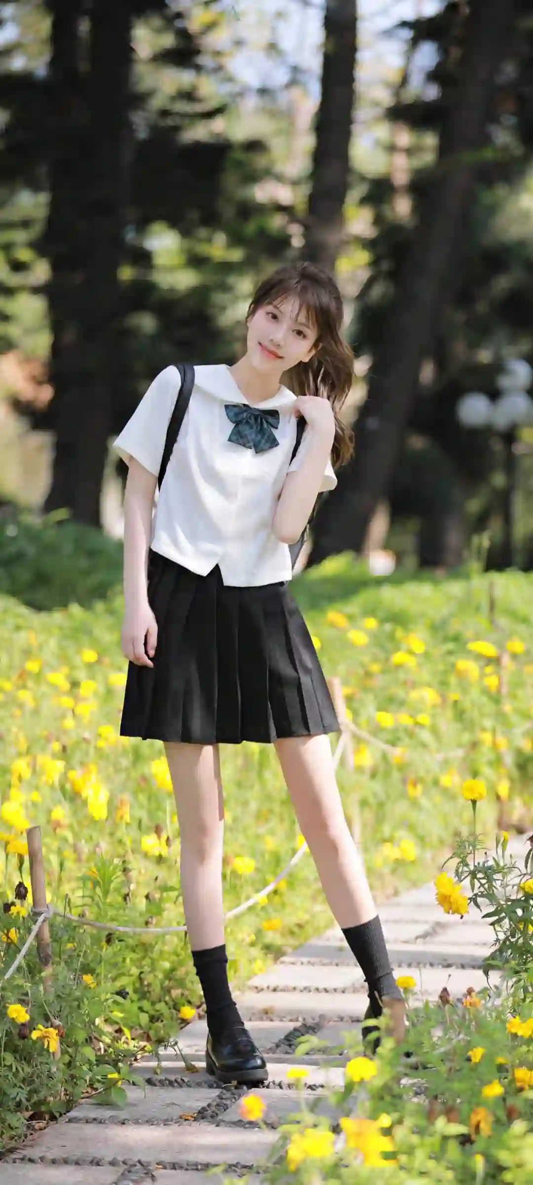 图片展示了一位穿着白色衬衫和黑色百褶裙的JK制服女孩 站在开满黄色花朵的户外小径上 背景是绿树和自然光 整体氛围清新自然