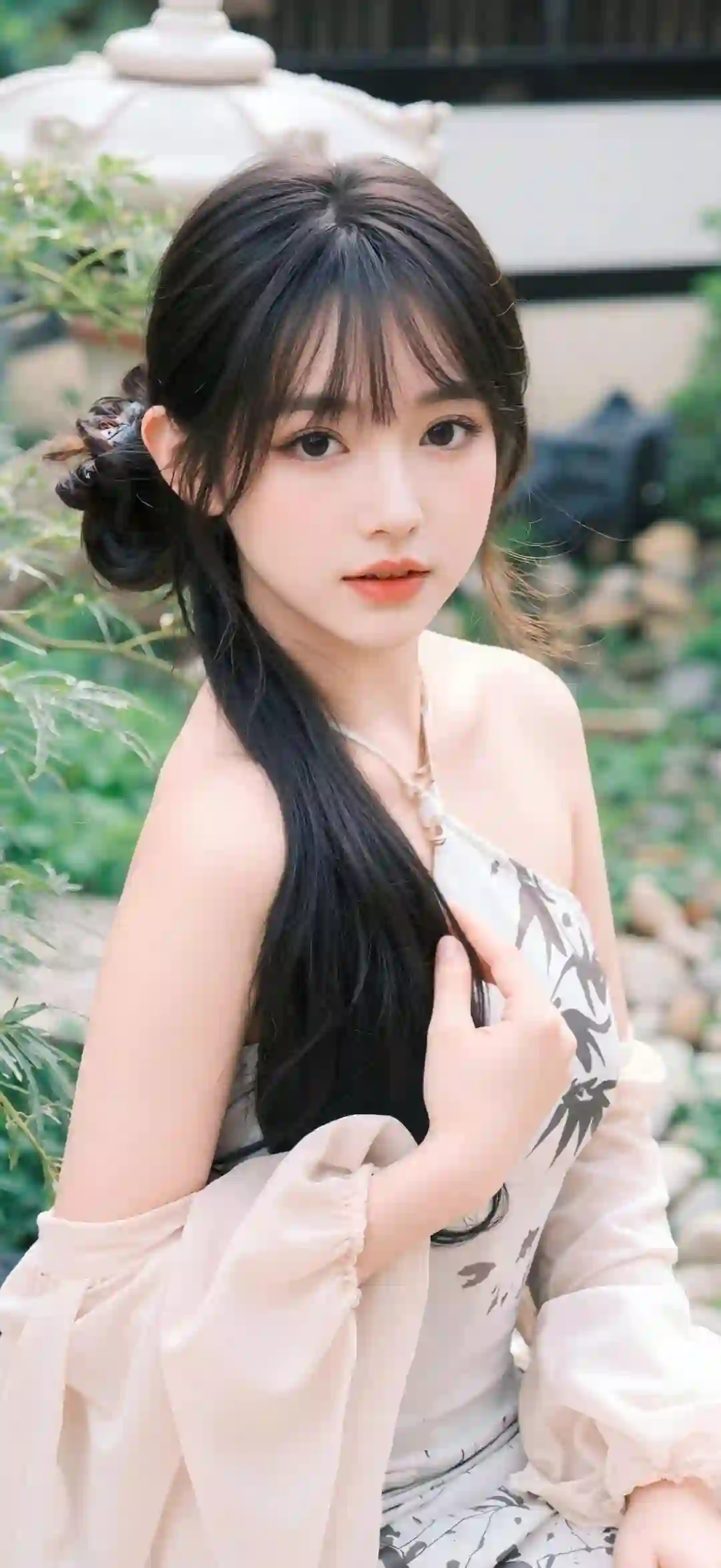 一位黑发年轻女性 穿着白色印花吊带裙和浅粉色薄纱罩衫 坐在户外庭院中 背景有绿植 表情温柔优雅 整体风格唯美清新