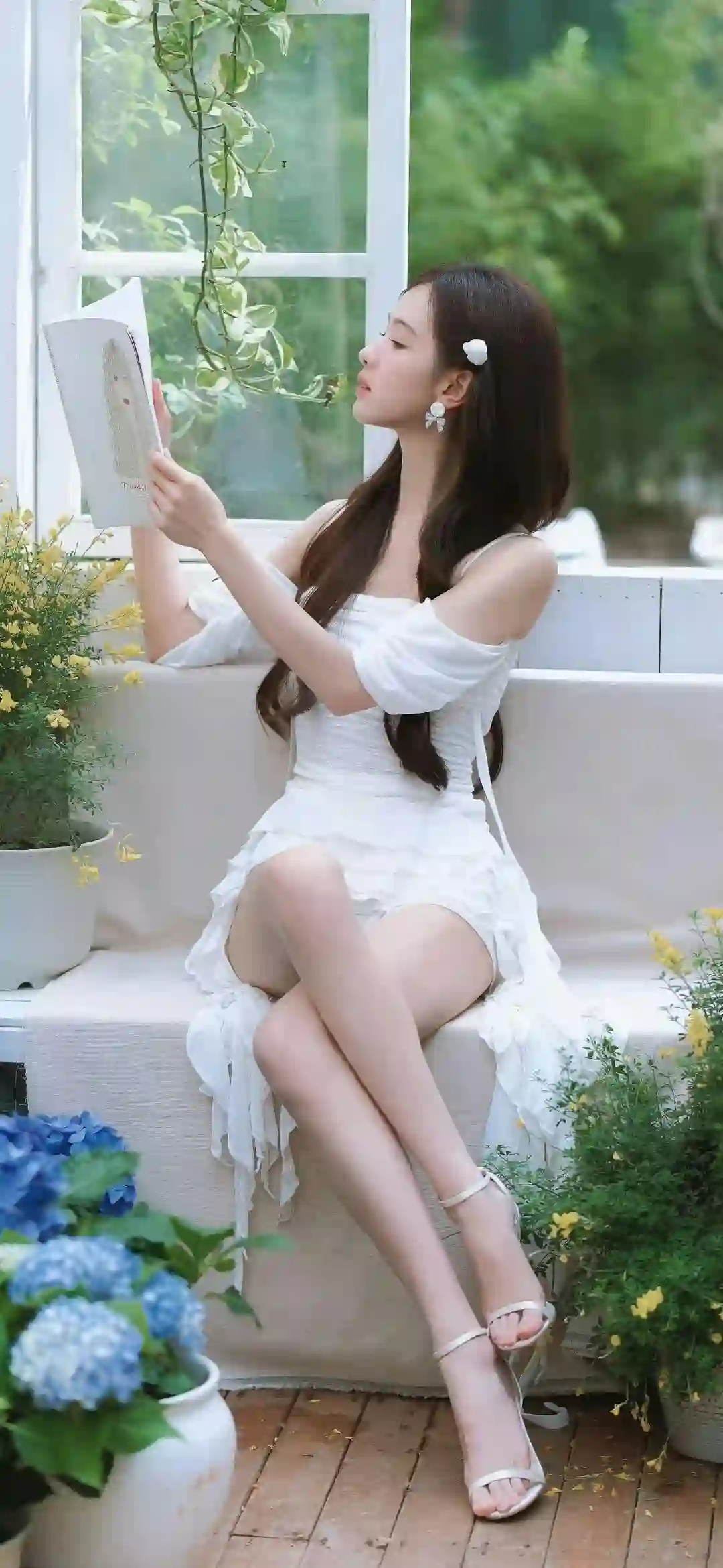一位穿着白色连衣裙的长发少女坐在窗边长椅上阅读 周围有蓝色绣球花和黄色小花 氛围清新优雅