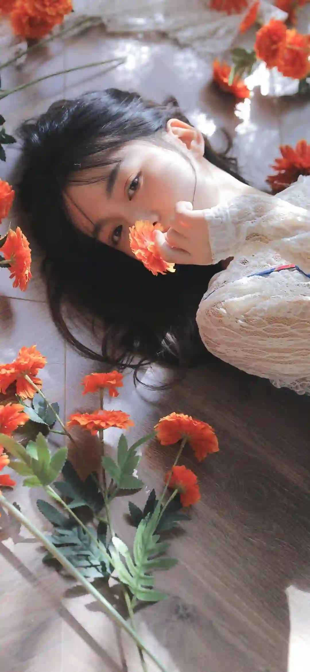 一位黑发女孩躺在木质地板上 手持橙色花朵 周围散落着橙色花朵 穿着白色蕾丝上衣 光线自然柔和 营造唯美氛围