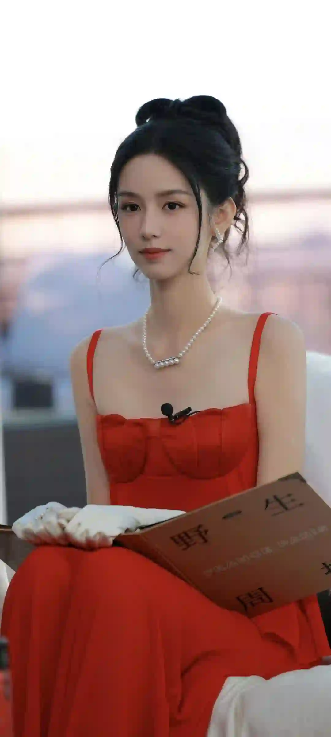 明星周也身穿红色吊带长裙 佩戴珍珠项链和白色耳环 黑色长发盘起 手戴白色手套 手持棕色封面书籍 整体造型优雅高贵 气质温婉动人