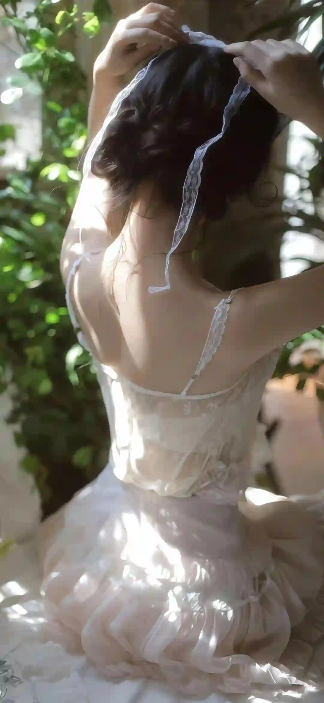 一位女孩背对镜头 穿着白色蕾丝裙 在阳光下整理白色头饰 背景有绿植 营造出唯美安静的氛围