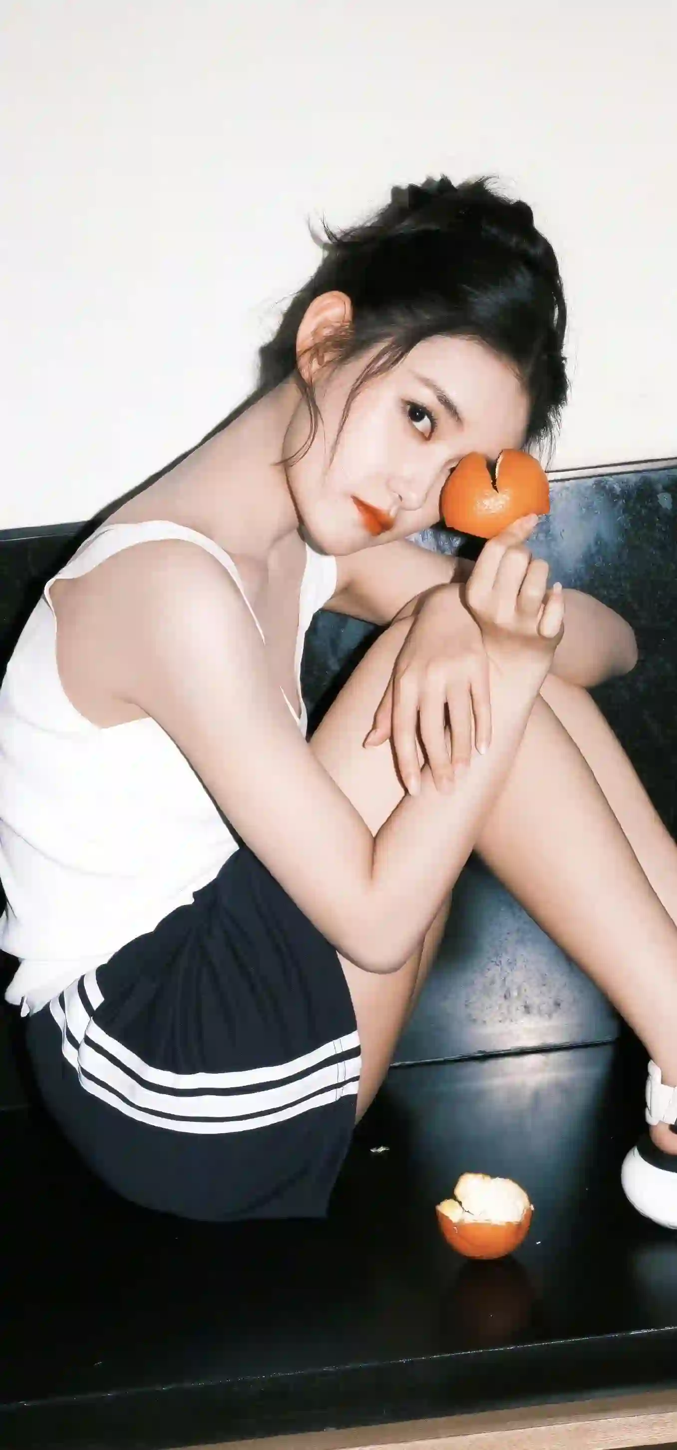 图片展示一位年轻女性 黑色长发盘起 身穿白色背心和黑色短裙 坐在黑色沙发上 手持橘子靠近脸颊 地面有橘子皮 整体氛围清新自然