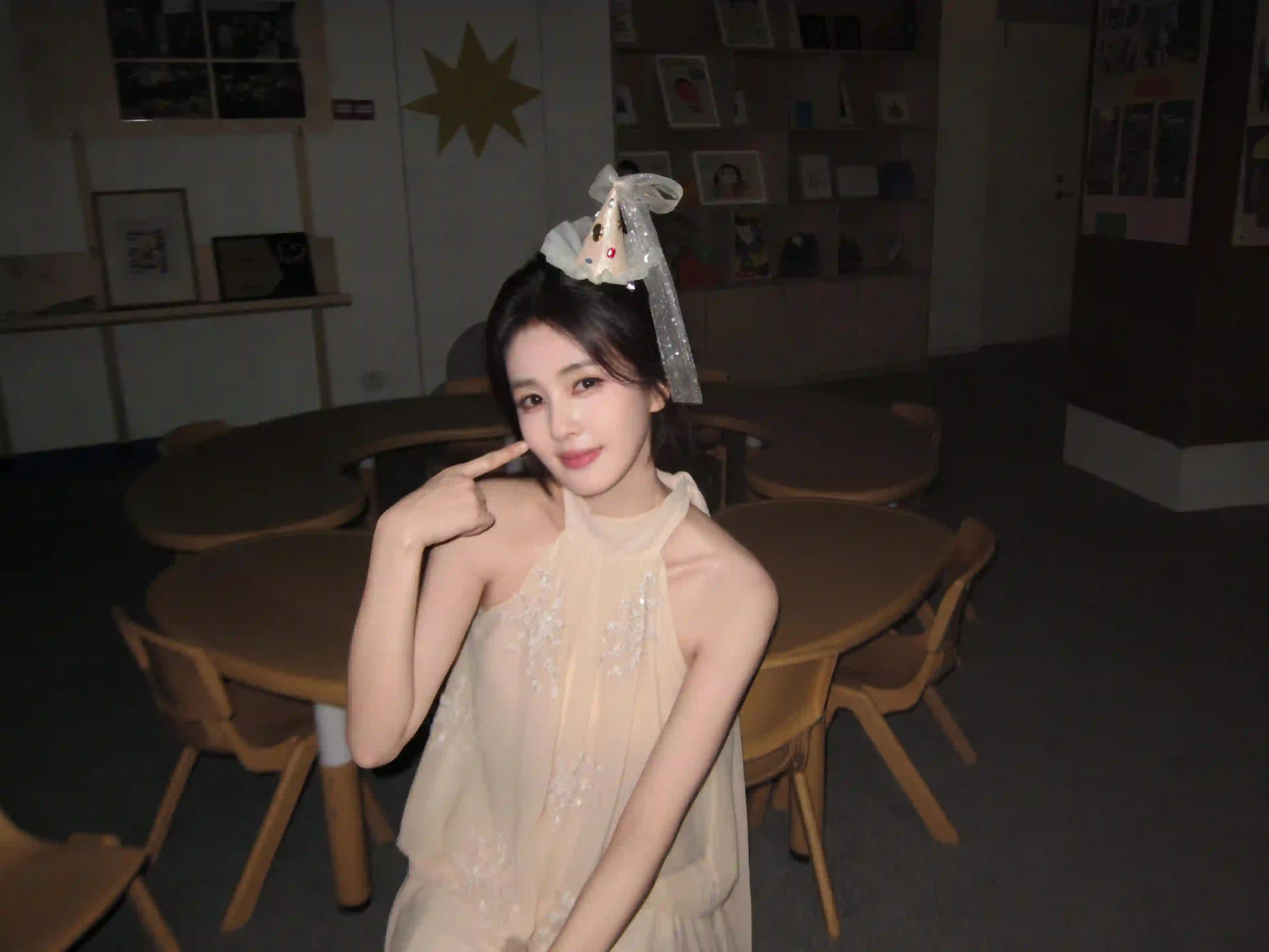 一位长发少女身穿米色连衣裙 头戴装饰性头饰 手轻触脸颊 背景是室内桌椅和架子 氛围温馨宁静