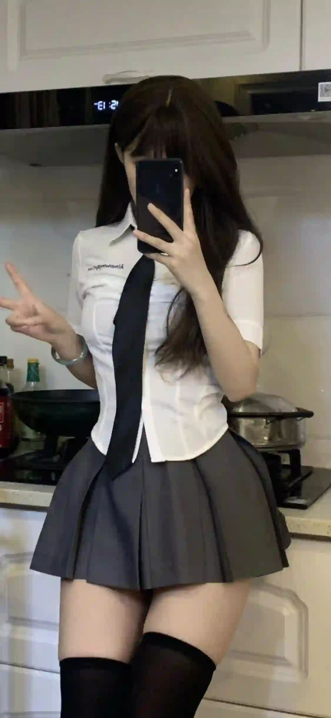 一位穿着白色衬衫 黑色领带和灰色百褶裙的年轻女性在厨房中用手机自拍 比出V手势 背景有锅具和调味品