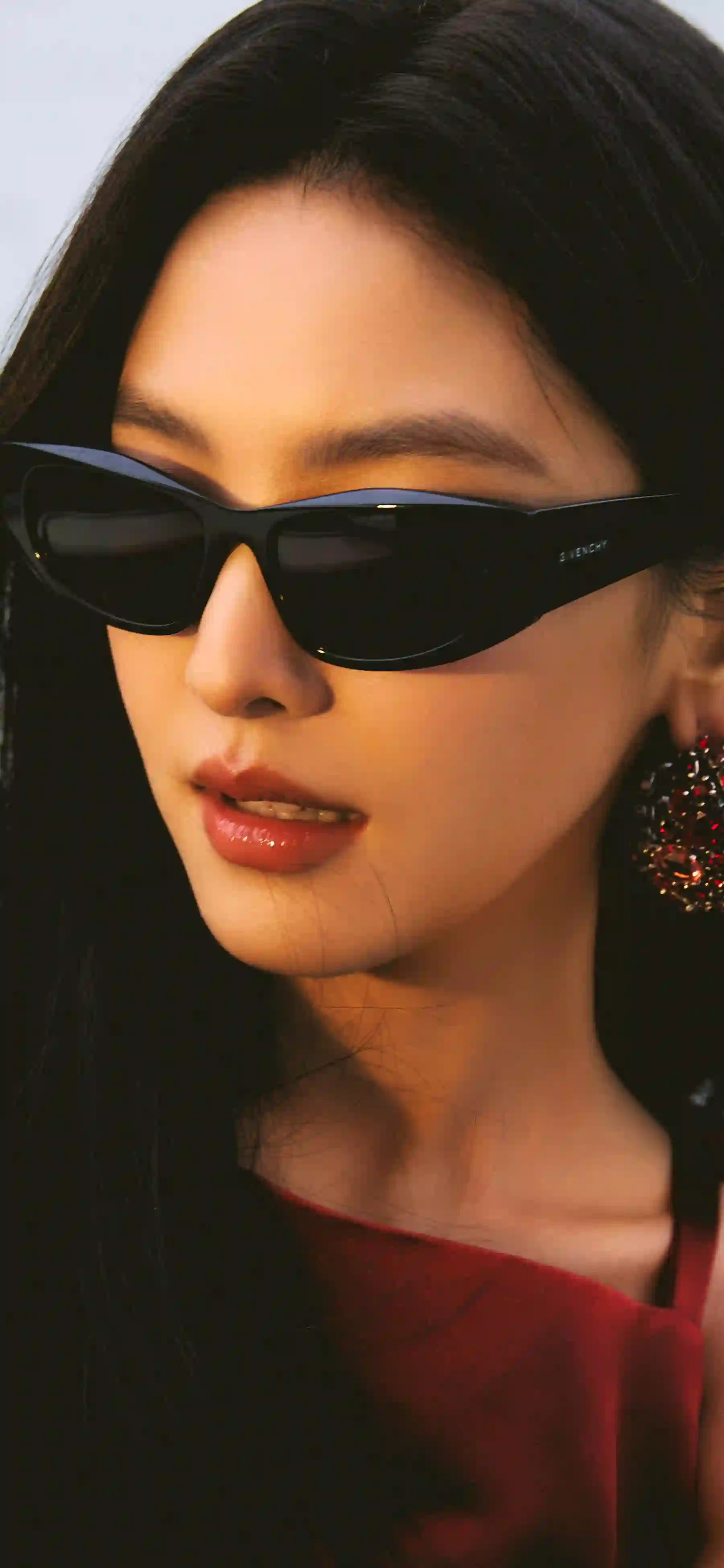 一位女性戴着黑色猫眼墨镜 穿着红色上衣 黑长直发 妆容精致 光线柔和 背景较暗 整体呈现时尚优雅的氛围