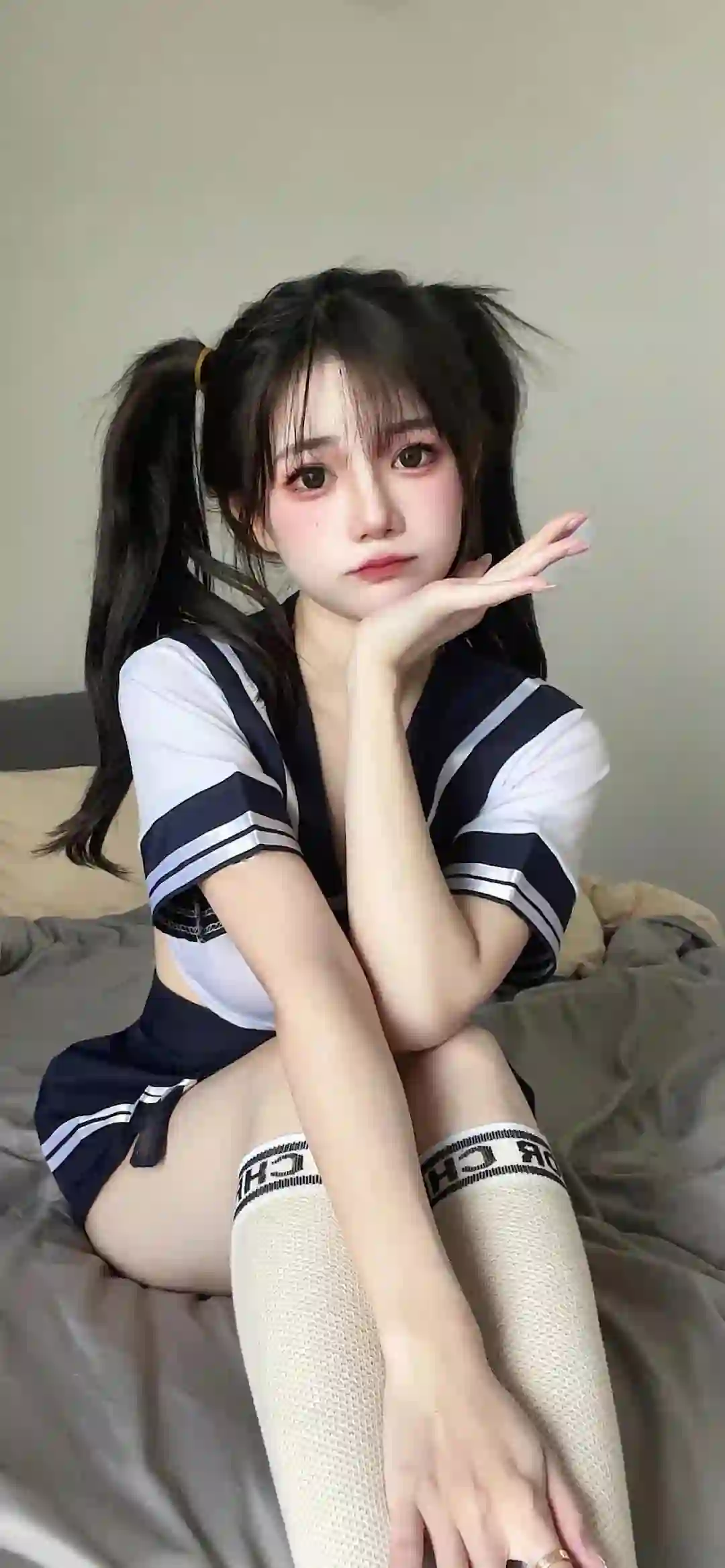 一位双马尾女孩穿着蓝白条纹校服，坐在床上优雅坐姿，手托下巴，展现清纯甜美气质。