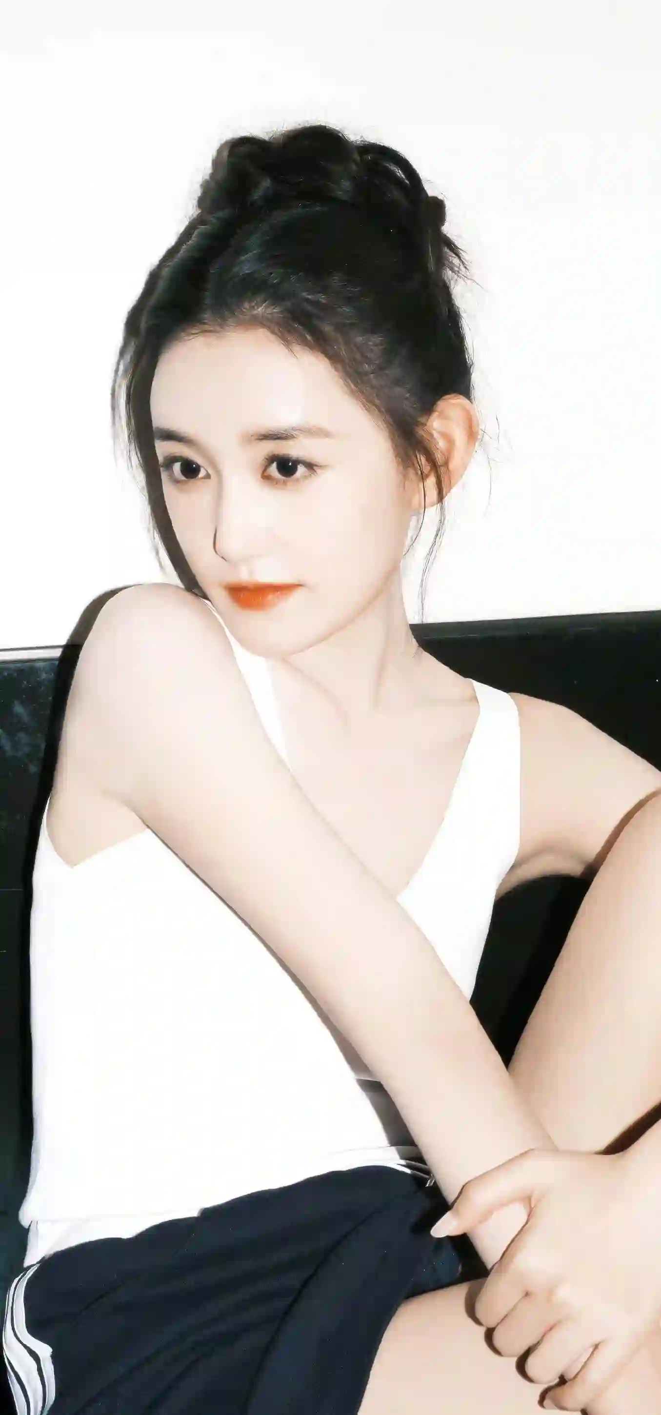一位黑色长发盘起的女性 穿着白色吊带上衣和深色裙子 坐在沙发上 姿势优雅 背景简洁