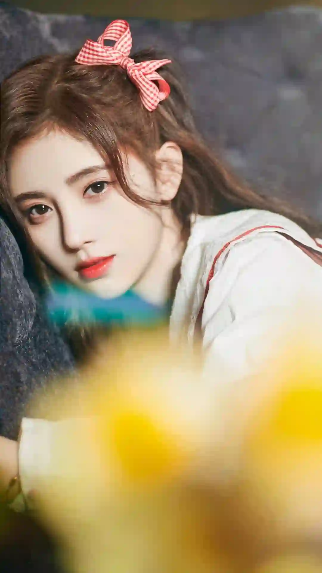 一位佩戴红白格子蝴蝶结发饰、妆容精致的年轻女性特写，侧靠在深色背景上，前景有模糊的黄色花朵，整体氛围温柔甜美。