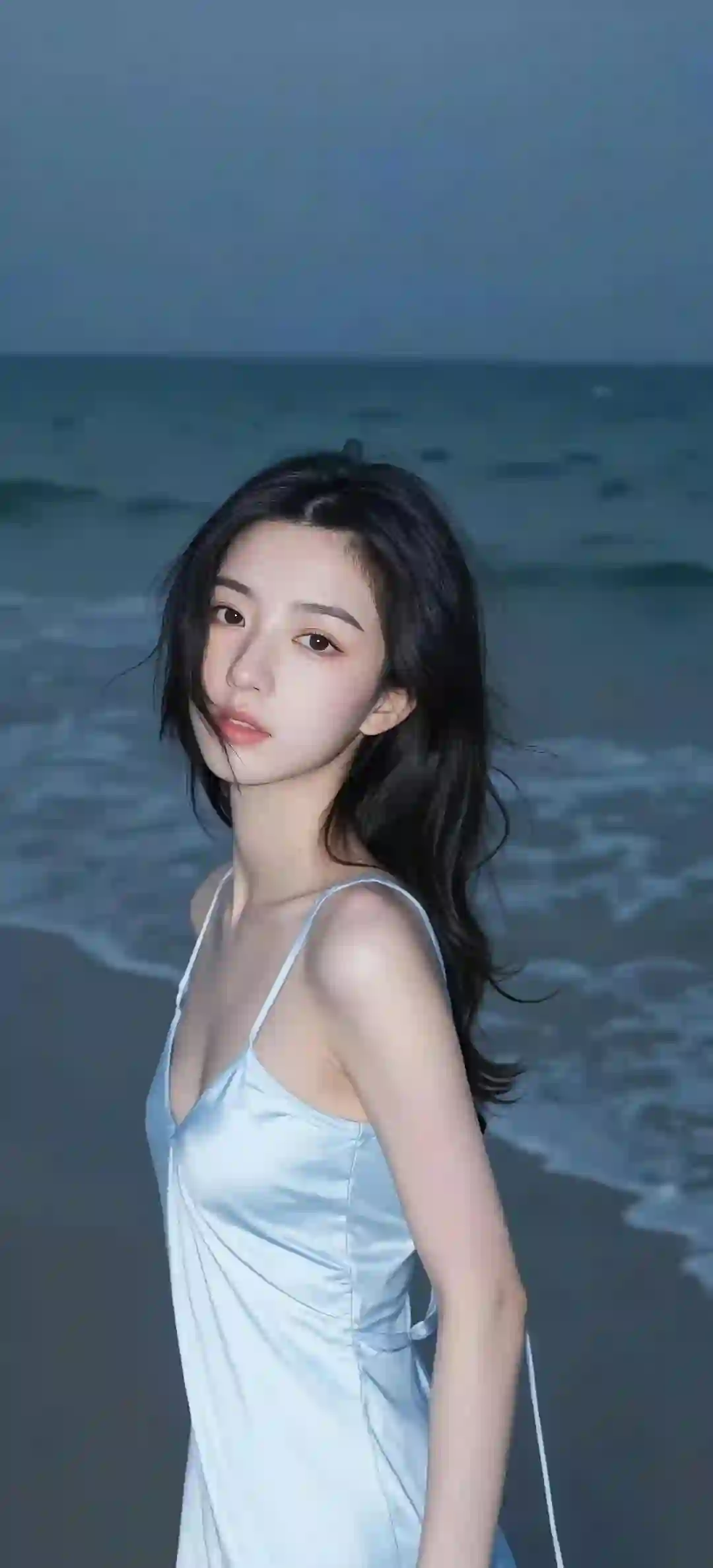 一位年轻女性身穿淡蓝色吊带连衣裙站在海边，黑发微卷，背景是蓝色大海和天空，整体氛围宁静唯美。
