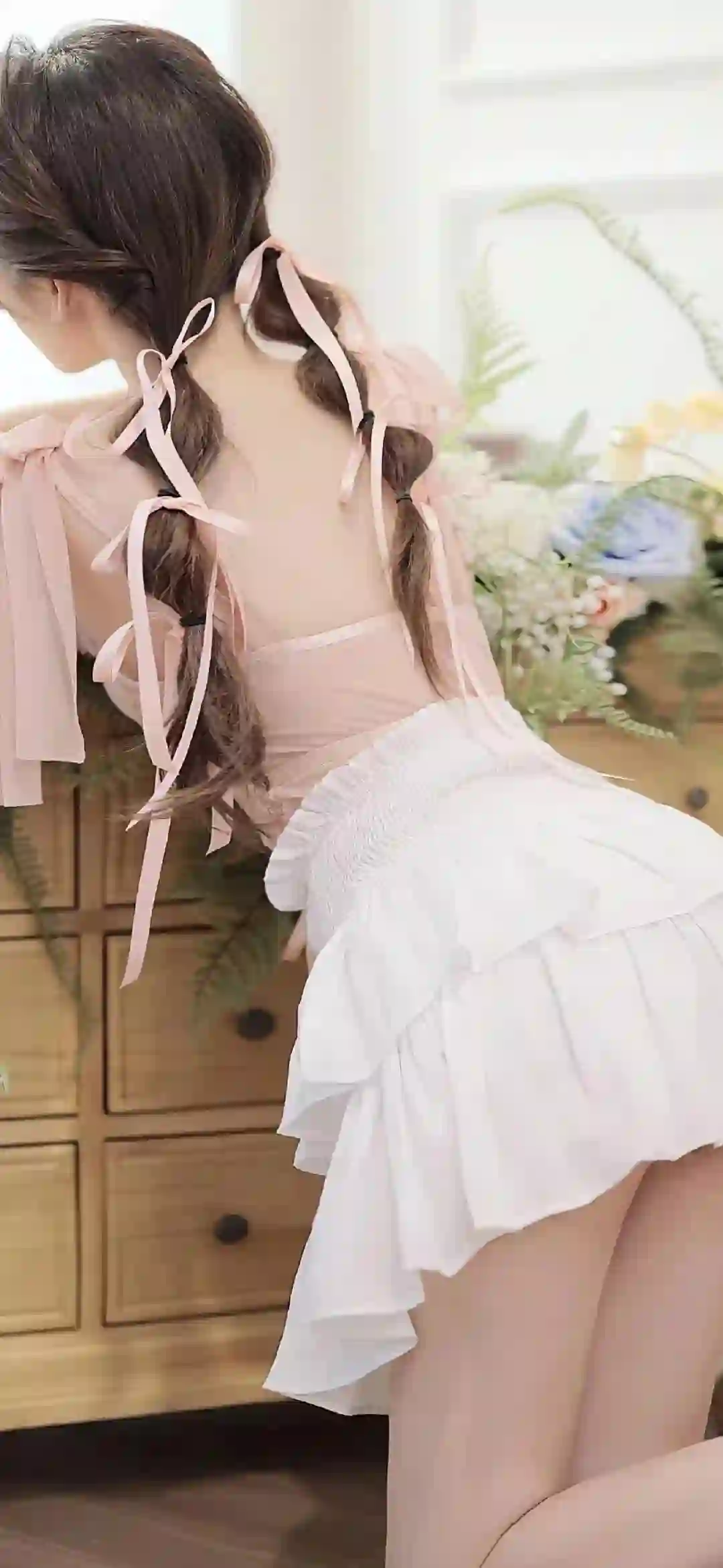 图片展示了一位背对镜头的少女 穿着粉色上衣和白色荷叶边裙子 头发编成双辫子并系有粉色丝带 背景是室内木质柜子和花卉装饰 整体风格清新甜美